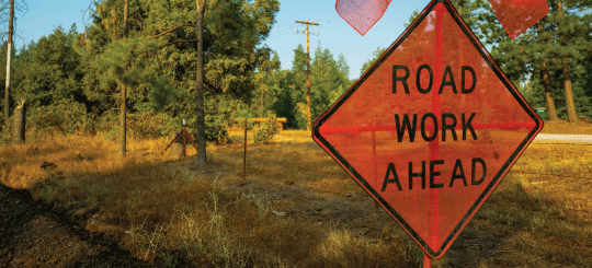 Orange Road Work Ahead firmar en un camino rural en un área natural con árboles y pastos en California