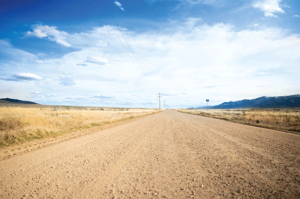 Fotografía de ángulo bajo de una carretera sin pavimentar tratada con Perma-Zyme en una pradera rural con montañas y postes de servicios públicos al fondo