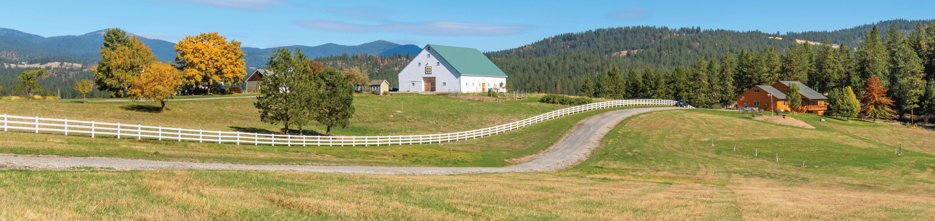 Propiedad agrícola residencial con camino de entrada, casa de troncos, granero blanco, valla blanca y montañas al fondo